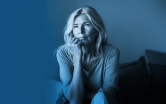 A thoughtful middle-aged woman with light hair sits indoors, resting her chin on her hand and gazing into the distance. The image has a blue tint, creating a calm and contemplative mood.