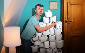 A man in a blue shirt stands surprised next to a tall stack of toilet paper rolls by a wooden door, holding several rolls in his arms. The scene is indoors with teal walls and a white lamp nearby.