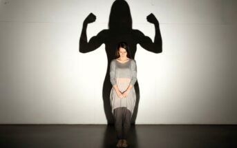 A woman stands barefoot with her head bowed, casting a shadow on the wall behind her that shows her in a strong, flexed-arm pose, symbolizing inner strength and confidence.